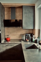 A contemporary kitchen featuring sleek gray cabinets and a countertop. The backsplash and cabinet exteriors are designed with a wood finish. On the counter, there is a flat stovetop with a red kettle, a coffee machine with containers, a glass, and a few other kitchen items. The sink is equipped with a metallic faucet, and there is a window providing natural light.
