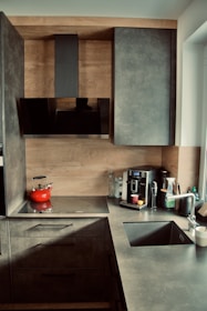 A contemporary kitchen featuring sleek gray cabinets and a countertop. The backsplash and cabinet exteriors are designed with a wood finish. On the counter, there is a flat stovetop with a red kettle, a coffee machine with containers, a glass, and a few other kitchen items. The sink is equipped with a metallic faucet, and there is a window providing natural light.