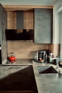 A contemporary kitchen featuring sleek gray cabinets and a countertop. The backsplash and cabinet exteriors are designed with a wood finish. On the counter, there is a flat stovetop with a red kettle, a coffee machine with containers, a glass, and a few other kitchen items. The sink is equipped with a metallic faucet, and there is a window providing natural light.