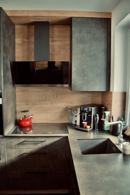 A contemporary kitchen featuring sleek gray cabinets and a countertop. The backsplash and cabinet exteriors are designed with a wood finish. On the counter, there is a flat stovetop with a red kettle, a coffee machine with containers, a glass, and a few other kitchen items. The sink is equipped with a metallic faucet, and there is a window providing natural light.