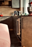 A modern kitchen area features sleek dark cabinets and a countertop with various items. Prominently placed is a red stand mixer, a black coffee maker, a red kettle, and several bottles and containers. The ambient lighting and neutral tones create a cozy atmosphere.