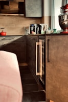 A modern kitchen area features sleek dark cabinets and a countertop with various items. Prominently placed is a red stand mixer, a black coffee maker, a red kettle, and several bottles and containers. The ambient lighting and neutral tones create a cozy atmosphere.