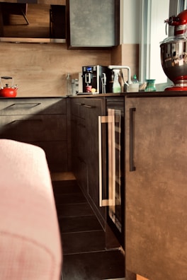 A modern kitchen area features sleek dark cabinets and a countertop with various items. Prominently placed is a red stand mixer, a black coffee maker, a red kettle, and several bottles and containers. The ambient lighting and neutral tones create a cozy atmosphere.