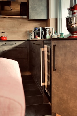 A modern kitchen area features sleek dark cabinets and a countertop with various items. Prominently placed is a red stand mixer, a black coffee maker, a red kettle, and several bottles and containers. The ambient lighting and neutral tones create a cozy atmosphere.