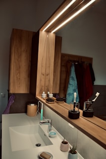 A bathroom counter featuring a modern design with a large mirror and wooden elements. The counter holds various items including an electric toothbrush, a bar of soap, small potted plants, and grooming accessories such as hairbrushes. The lighting is contemporary with strip lights above the mirror, enhancing the minimalist aesthetic.