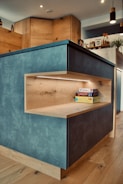 A modern kitchen with wood-paneled cabinets and a blue-grey island. The island features a built-in wooden shelf illuminated by LED lighting, holding some board games. Various kitchen items, plants, and decorations are visible on the countertop and shelves in the background.