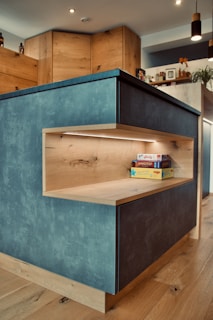 A modern kitchen with wood-paneled cabinets and a blue-grey island. The island features a built-in wooden shelf illuminated by LED lighting, holding some board games. Various kitchen items, plants, and decorations are visible on the countertop and shelves in the background.
