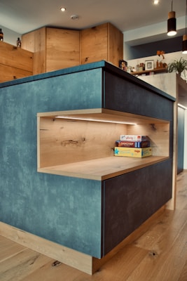 A modern kitchen with wood-paneled cabinets and a blue-grey island. The island features a built-in wooden shelf illuminated by LED lighting, holding some board games. Various kitchen items, plants, and decorations are visible on the countertop and shelves in the background.