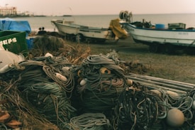 Bundles of thick ropes and fishing equipment lie on a sandy beach in close proximity to two fishing boats. The scene is set near a calm sea, with a slightly overcast sky and a generally muted color palette. The ropes are tangled and interwoven with buoys and other maritime tools, casting a sense of disuse or preparation for future use.