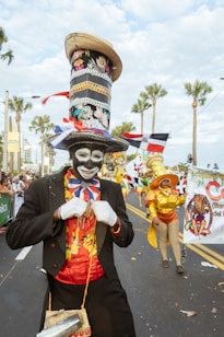 A person in elaborate costume stands in the forefront, wearing a tall, decorative hat with patterns and a face painted in white with striking features. Others in equally vibrant attire are in the background. The scene is lively, indicating a festive atmosphere on a street lined with palm trees under a partly cloudy sky.