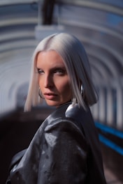 A woman with platinum blonde hair, styled in a sleek bob, gazes intently at the camera. She is wearing a shiny dark jacket and large hoop earrings. The background consists of an arched structure with a mix of shadows and light, creating a tunnel-like effect.