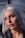 A close-up portrait of a person with platinum blonde hair, soft makeup, and a neutral expression, captured in soft lighting with a blurred background.