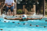 A swimmer is captured mid-stroke in a pool, demonstrating the butterfly technique with arms outstretched. Water splashes around, indicating motion and energy. The swimmer is wearing a cap and goggles. In the background, there is a person seated, observing the activity, and out of focus greenery and structures.