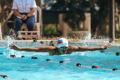 A swimmer is captured mid-stroke in a pool, demonstrating the butterfly technique with arms outstretched. Water splashes around, indicating motion and energy. The swimmer is wearing a cap and goggles. In the background, there is a person seated, observing the activity, and out of focus greenery and structures.