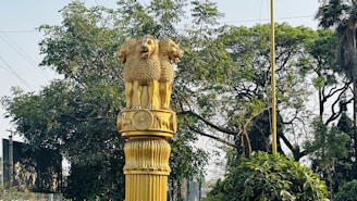A golden statue featuring the Ashoka Pillar lion capital symbol stands prominently against a backdrop of lush green trees and foliage. The sculpture is intricately designed with three lions and is mounted on a textured column. The scene conveys a sense of national significance and artistic craftsmanship.