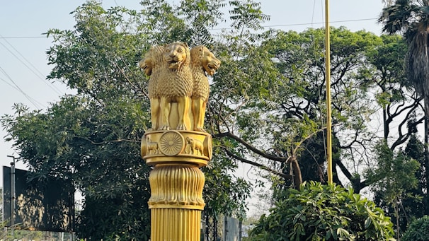 A golden statue featuring the Ashoka Pillar lion capital symbol stands prominently against a backdrop of lush green trees and foliage. The sculpture is intricately designed with three lions and is mounted on a textured column. The scene conveys a sense of national significance and artistic craftsmanship.