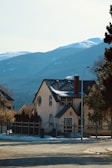 A quaint house with a sloped roof and chimney, set against a backdrop of snow-capped mountains. The house is surrounded by trees and has a fenced yard. The scene suggests a peaceful residential area with clear skies and sunlight illuminating the facade.
