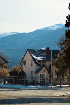 A quaint house with a sloped roof and chimney, set against a backdrop of snow-capped mountains. The house is surrounded by trees and has a fenced yard. The scene suggests a peaceful residential area with clear skies and sunlight illuminating the facade.