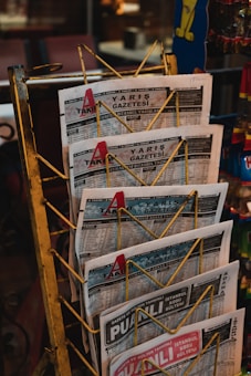 A rack holds multiple copies of a newspaper with the title 'Yarış Gazetesi'. The newspapers are arranged neatly in a yellow metal holder. The setting appears to be a storefront or kiosk, with other items partially visible in the background.