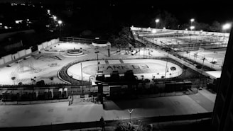 A nighttime aerial view of an outdoor recreational facility with various sports and play areas. The foreground features a running track with the words 'PARK G3' written on it. Surrounding the track are playground structures and a round seating area. Further in the background, several tennis courts and a basketball court are visible, with bright lights illuminating the entire area.