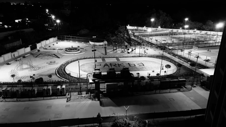 A nighttime aerial view of an outdoor recreational facility with various sports and play areas. The foreground features a running track with the words 'PARK G3' written on it. Surrounding the track are playground structures and a round seating area. Further in the background, several tennis courts and a basketball court are visible, with bright lights illuminating the entire area.
