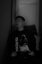 A person is sitting against a wall, wearing a dark sweater with the word 'vedanta' and an image printed on it. The setting appears to be indoors with subdued lighting, and a stack of chairs is visible in the background.