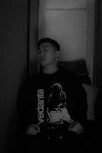 A person is sitting against a wall, wearing a dark sweater with the word 'vedanta' and an image printed on it. The setting appears to be indoors with subdued lighting, and a stack of chairs is visible in the background.