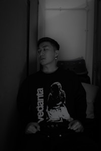 A person is sitting against a wall, wearing a dark sweater with the word 'vedanta' and an image printed on it. The setting appears to be indoors with subdued lighting, and a stack of chairs is visible in the background.