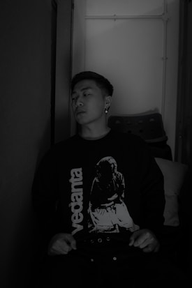 A person is sitting against a wall, wearing a dark sweater with the word 'vedanta' and an image printed on it. The setting appears to be indoors with subdued lighting, and a stack of chairs is visible in the background.