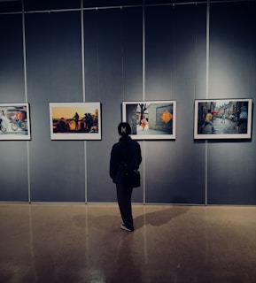 A person stands in front of a gallery wall, observing four framed photographs. The room has dim lighting, creating a contemplative atmosphere as the individual looks at the art.
