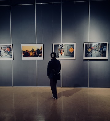 A person stands in front of a gallery wall, observing four framed photographs. The room has dim lighting, creating a contemplative atmosphere as the individual looks at the art.