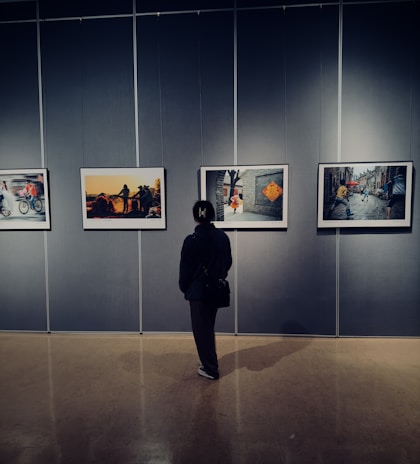 A person stands in front of a gallery wall, observing four framed photographs. The room has dim lighting, creating a contemplative atmosphere as the individual looks at the art.