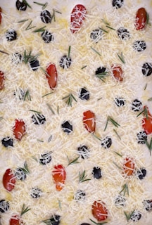 A flat surface covered with shredded cheese, scattered black olives, cherry tomatoes halved, and sprigs of rosemary arranged in an artistic manner, resembling a pizza or focaccia topping.