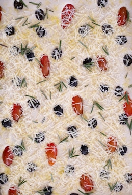 A flat surface covered with shredded cheese, scattered black olives, cherry tomatoes halved, and sprigs of rosemary arranged in an artistic manner, resembling a pizza or focaccia topping.