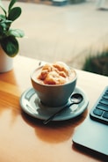 A cup of coffee with ice cream on top sits on a saucer beside a spoon and a laptop. A small potted plant adds a touch of greenery on the wooden table by the window.