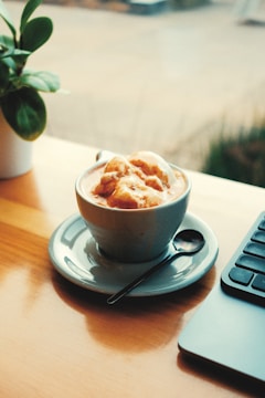 A cup of coffee with ice cream on top sits on a saucer beside a spoon and a laptop. A small potted plant adds a touch of greenery on the wooden table by the window.