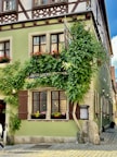 A picturesque corner house with traditional architectural style featuring wooden accents and lush green ivy climbing up the walls. The building has multiple windows adorned with red flower boxes, and the ground floor features a sign indicating it's a hotel. The house is painted a light green color, and there are cobblestone streets surrounding it.