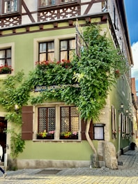 A picturesque corner house with traditional architectural style featuring wooden accents and lush green ivy climbing up the walls. The building has multiple windows adorned with red flower boxes, and the ground floor features a sign indicating it's a hotel. The house is painted a light green color, and there are cobblestone streets surrounding it.