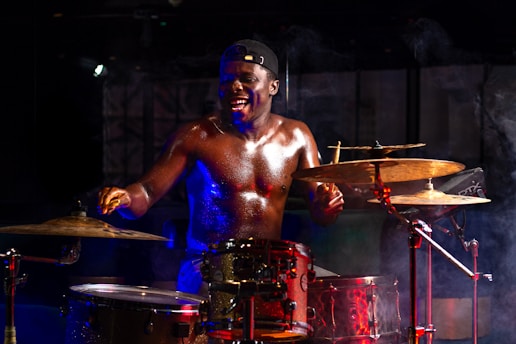 A shirtless man energetically playing drums with a wide smile, captured amidst dynamic stage lighting and smoke effects. He wears a backwards cap and is surrounded by an array of cymbals and drums.