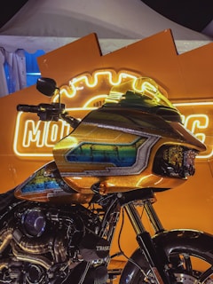 A motorcycle with a custom paint job featuring vibrant blue and green accents is positioned in front of a bright orange backdrop with illuminated signage. The signage has a neon glow, adding to the striking contrast with the motorcycle's design. The scene appears to be a display under a tent, possibly at a show or exhibition.