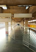 A deserted railway platform with concrete pillars and metal railings. The platform is well-lit with natural light coming from openings at the top. The space appears expansive due to the linear perspective and repeating patterns of the structure.