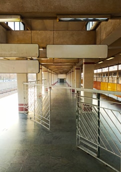 A deserted railway platform with concrete pillars and metal railings. The platform is well-lit with natural light coming from openings at the top. The space appears expansive due to the linear perspective and repeating patterns of the structure.
