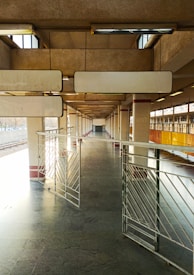 A deserted railway platform with concrete pillars and metal railings. The platform is well-lit with natural light coming from openings at the top. The space appears expansive due to the linear perspective and repeating patterns of the structure.