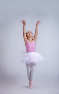 A young ballerina wearing a pink leotard and white tutu is standing on pointed toes with arms gracefully extended upwards. The background is a plain, light gray, adding emphasis to the dancer.