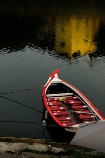 A small boat with a black and red exterior is tied to the shore. The boat is equipped with several red seating cushions arranged in rows. The water reflects the silhouette of a yellow structure or object.