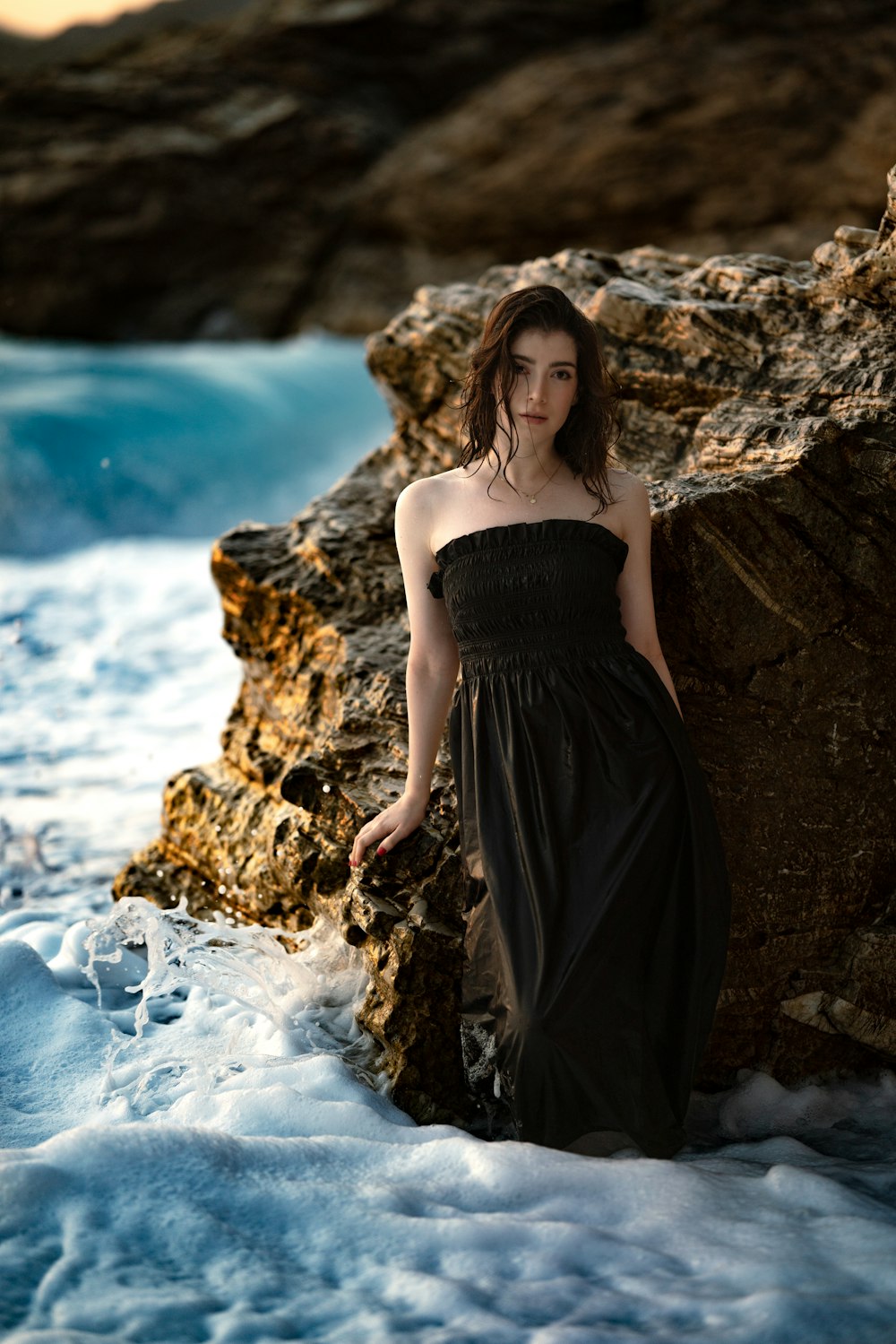 a woman in a black dress standing on a rock by the ocean