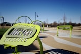 Bright green outdoor musical instruments are set up in a public park. The instruments, which have a modern design, are situated on a paved area, surrounded by green grass and trees. In the background, a clear blue sky and additional park features like pathways and fences are visible.