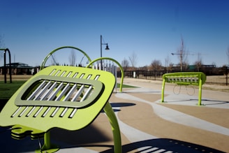Bright green outdoor musical instruments are set up in a public park. The instruments, which have a modern design, are situated on a paved area, surrounded by green grass and trees. In the background, a clear blue sky and additional park features like pathways and fences are visible.