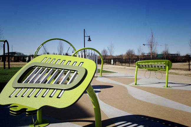 Bright green outdoor musical instruments are set up in a public park. The instruments, which have a modern design, are situated on a paved area, surrounded by green grass and trees. In the background, a clear blue sky and additional park features like pathways and fences are visible.