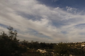 A landscape featuring a wide, open sky with scattered clouds and a view of rolling hills. Some vegetation and trees can be seen in the foreground, adding texture to the expansive view. The image has a slightly blurred effect, suggesting motion.